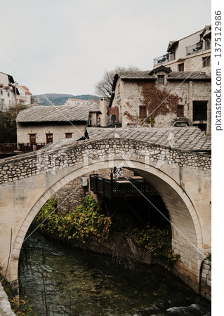 Kriva Cuprija stone bridge in Mostar crossing a shallow river, surrounded by historic houses and old town architecture. Medieval Balkan landmark reflecting cultural heritage and calm urban atmosphere 137512986