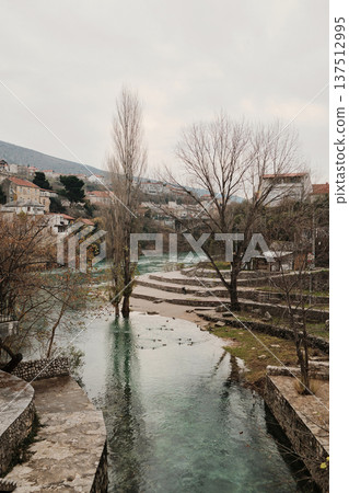 Calm section of the Neretva River in Mostar with trees, stone embankments, and city houses along the water. Peaceful urban landscape combining nature and historic surroundings Calm section of the Neretva River in Mostar with trees, stone embankments, and city houses along the water. Peaceful urban landscape combining nature and historic surroundings 137512995