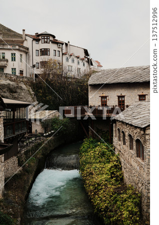 Narrow river canal flowing through the old town of Mostar between stone houses and wooden terraces. Quiet historic city scene showing traditional Balkan architecture and everyday atmosphere Narrow river canal flowing through the old town of Mostar between stone houses and wooden terraces. Quiet historic city scene showing traditional Balkan architecture and everyday atmosphere 137512996