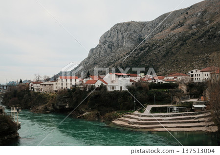 Residential area of Mostar along the turquoise Neretva River with stone terraces, houses, and steep mountain slopes. Calm Balkan city landscape showing everyday life and natural surroundings 137513004