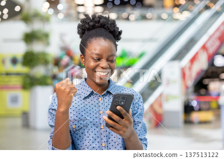 Happy young woman in a busy shopping mall celebrates good news on her smartphone, smiling and pumping a fist in triumph while browsing stores and using a mobile app Happy young woman in a busy shopping mall celebrates good news on her smartphone, smiling and pumping a fist in triumph while browsing stores and using a mobile app 137513122