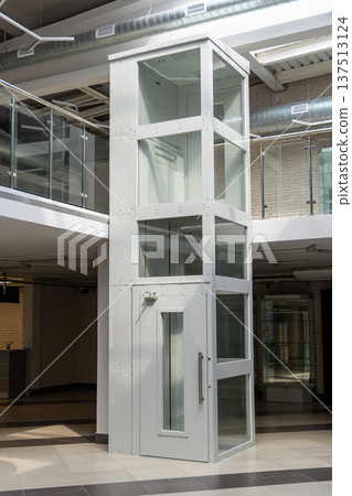 Modern glass elevator in a bright, industrial-style lobby. Modern glass elevator in a bright, industrial-style lobby. 137513124