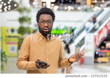 Confused individual checking phone and credit card, experiencing an unexpected financial issue or fraud while standing bewildered in a busy shopping center 137513135