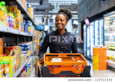 Young woman smiling while pushing an orange shopping cart down a supermarket aisle, choosing fresh groceries and enjoying a casual, everyday shopping trip for daily needs 137513143