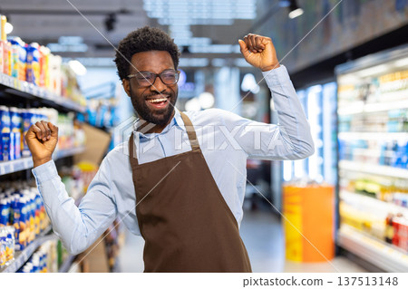 Energetic shop assistant in apron and glasses dances with raised arms and broad smile between grocery store shelves, celebrating success, joy and confident customer service 137513148