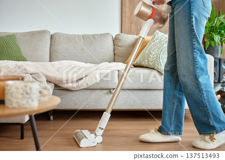 Woman vacuuming floor with cordless vacuum cleaner in home interior near sofa Woman vacuuming floor with cordless vacuum cleaner in home interior near sofa 137513493