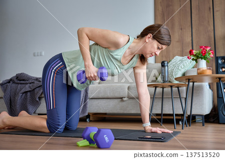 Woman performs one arm dumbbell row on exercise mat in living room with smartphone nearby Woman performs one arm dumbbell row on exercise mat in living room with smartphone nearby 137513520
