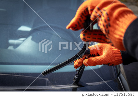 Man inspects windshield wipers on a car while wearing bright gloves in clear daylight outside Man inspects windshield wipers on a car while wearing bright gloves in clear daylight outside 137513585
