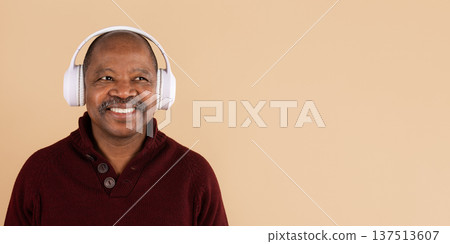 Smiling black man wearing white headphones listening to music on beige background Smiling black man wearing white headphones listening to music on beige background 137513607