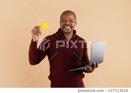 Mature black man holding credit card and laptop on beige studio background Mature black man holding credit card and laptop on beige studio background 137513628