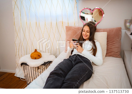 Teenager relaxing in bedroom smiling and texting on smartphone Teenager relaxing in bedroom smiling and texting on smartphone 137513730