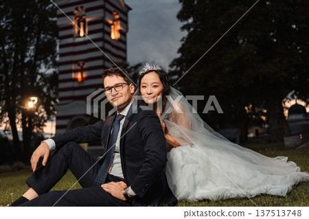 Interracial wedding couple sitting on grass at dusk Interracial wedding couple sitting on grass at dusk 137513748