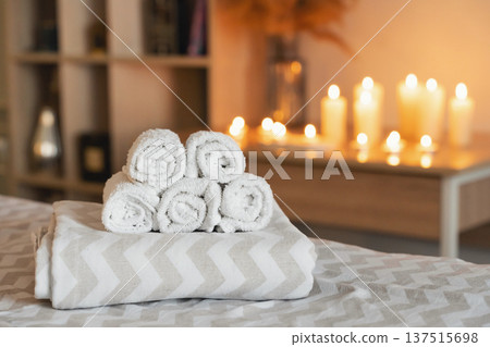 Rolled white towels stacked on a bed or massage table, offering a concept of spa, wellness, and relaxation with blurred candles 137515698