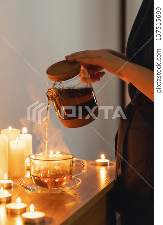 Woman pouring hot herbal tea into a glass cup with steam rising, surrounded by glowing candles, creating a moment of comfort and wellbeing Woman pouring hot herbal tea into a glass cup with steam rising, surrounded by glowing candles, creating a moment of comfort and wellbeing 137515699