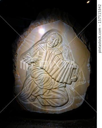 Bas-Relief Sculpture of a Woman Playing an Accordion. Bas-relief sculpture featuring a woman playing an accordion, intricately carved into a polished stone surface. 137515842