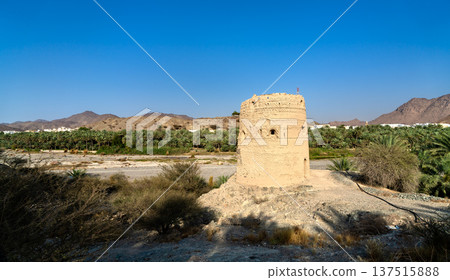 The historic stone structure of Al Basra Fort Tower stands near a lush palm oasis and dry riverbed in the scenic mountain landscape of Fanja, Oman. 137515888