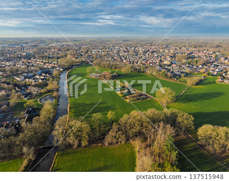 Wide aerial panorama of a Dutch town border with a winding canal, green meadows, and residential districts. Scenic European rural urban landscape under a blue sky at sunset. Wide aerial panorama of a Dutch town border with a winding canal, green meadows, and residential districts. Scenic European rural urban landscape under a blue sky at sunset. 137515948