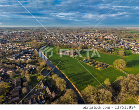 High aerial panorama of a Dutch town with a winding canal, green fields, and suburban housing. Scenic rural urban landscape in the Netherlands under a cloudy blue sky at golden hour. High aerial panorama of a Dutch town with a winding canal, green fields, and suburban housing. Scenic rural urban landscape in the Netherlands under a cloudy blue sky at golden hour. 137515954