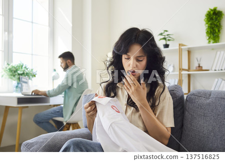 Jealous wife in shock and stress holding husbands shirt with lipstick marks 137516825