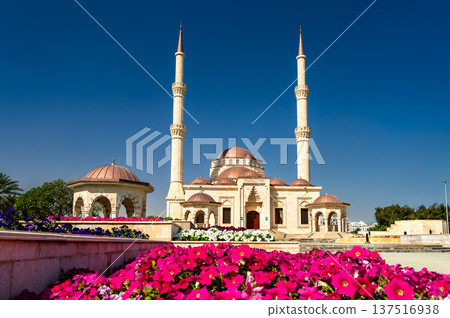 Architectural view of the Said Bin Taimur Mosque in Muscat, Oman, featuring twin minarets, domes, and colorful flower beds in the foreground under a clear blue sky. 137516938