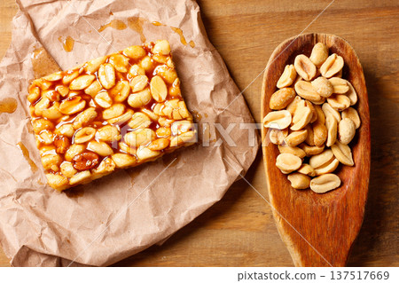 A piece of Brazilian peanut brittle made with roasted peanuts and golden caramel, on kraft paper on a rustic wooden table next to a spoon A piece of Brazilian peanut brittle made with roasted peanuts and golden caramel, on kraft paper on a rustic wooden table next to a spoon 137517669