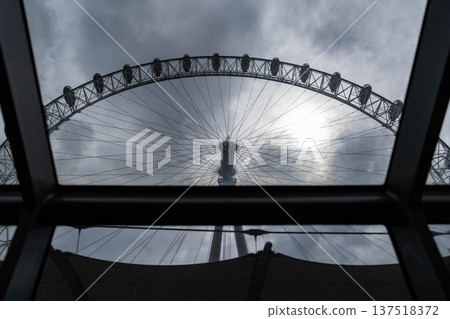 Upward view from inside a glass passenger cabin toward the central hub and cable structure of a large observation wheel framed by window glass against a cloudy sky. Upward view from inside a glass passenger cabin toward the central hub and cable structure of a large observation wheel framed by window glass against a cloudy sky. 137518372