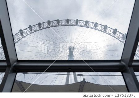 Upward view from inside a glass passenger cabin toward the central hub and cable structure of a large observation wheel framed by window glass against a cloudy sky. Upward view from inside a glass passenger cabin toward the central hub and cable structure of a large observation wheel framed by window glass against a cloudy sky. 137518373