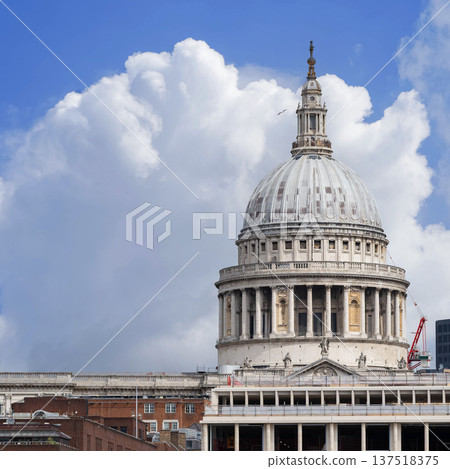 St Paul Cathedral dome rises above the River Thames in London with Millennium Bridge leading toward the historic landmark under a dramatic cloudy sky. 137518375