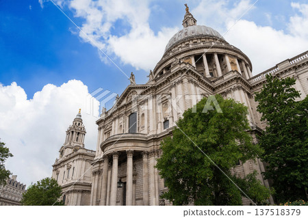 Historic architecture of St Paul Cathedral in London rises behind Millennium Bridge with pedestrians in the foreground beneath dramatic clouds in the sky. 137518379