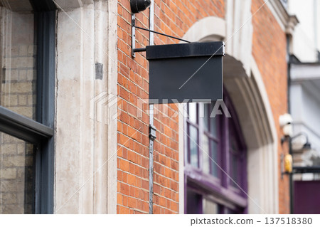 Blank metal shop sign hangs from a brick wall on a traditional street building in England with empty space for business branding or store name. Blank metal shop sign hangs from a brick wall on a traditional street building in England with empty space for business branding or store name. 137518380