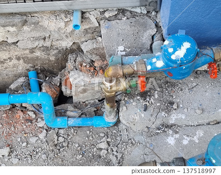 Blue water meter and PVC pipes with brass fittings installed against a damaged concrete wall. Residential plumbing infrastructure and utility equipment on a rough industrial construction background. Blue water meter and PVC pipes with brass fittings installed against a damaged concrete wall. Residential plumbing infrastructure and utility equipment on a rough industrial construction background. 137518997