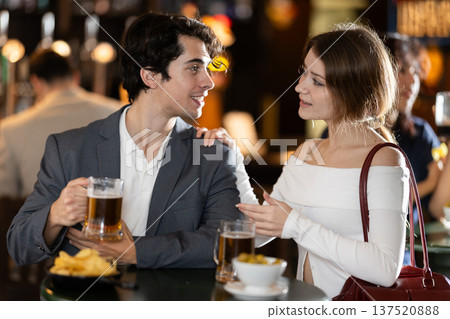 Young positive couple drinking and chatting while sitting at table in cozy bar Young positive couple drinking and chatting while sitting at table in cozy bar 137520888