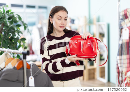 Fashionable young female client standing against background of shelves with clothes in store and choosing handbag. Shopping Christmas and New Year sales 137520962