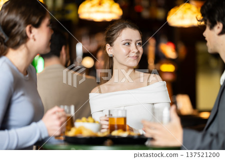 Group of positive young friends talking cheerfully and drinking beer at table in bar 137521200