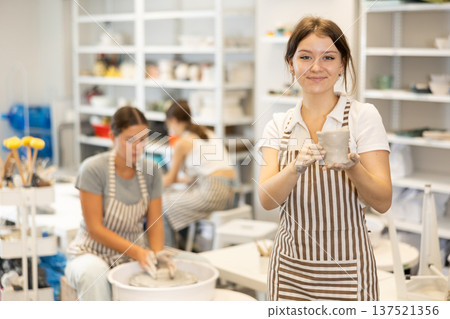 Girl takes break, stands and rests, waves hello with her hands stained with clay during pottery class Girl takes break, stands and rests, waves hello with her hands stained with clay during pottery class 137521356