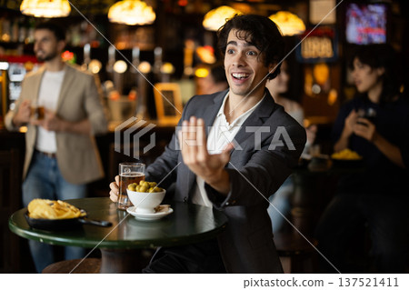 Positive young guy relaxing at table, drinking beer in sports pub and waiting for friends, raising hand in greeting 137521411