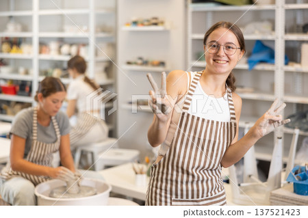 Teen girl takes break, rests, waves hello with her hands stained with clay during pottery class Teen girl takes break, rests, waves hello with her hands stained with clay during pottery class 137521423