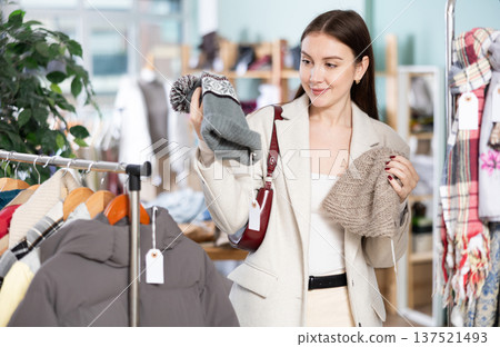 Focused european young woman choose hat on sale of winter clothing collection in clothing store Focused european young woman choose hat on sale of winter clothing collection in clothing store 137521493