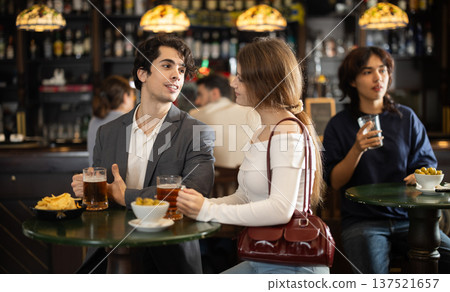 Couple of young guy and girl chatting, drinking beer and eating snacks at table in sports pub 137521657