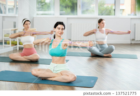 Girl conducts Pilates class for girls sitting with elastic band in hands above head makes body bends 137521775