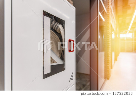 Fire safety equipment in a modern office. White hydrant cabinet with a fire hose behind glass in an industrial interior corridor. 137522033