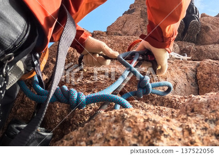 Close-up of mountain climber hands in gloves securing a blue safety rope with knots on a steep rock face during an ascent. Close-up of mountain climber hands in gloves securing a blue safety rope with knots on a steep rock face during an ascent. 137522056