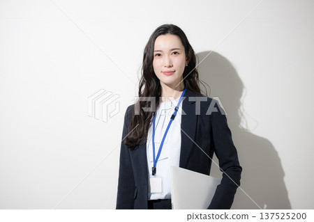 一位身著職業套裝、佩戴名牌、手持筆記型電腦的幹練女性，站在白色背景前。畫面中，她正抬頭展望未來和目標。 137525200