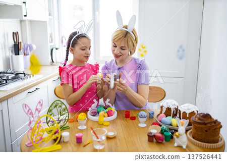 A mother and daughter wearing bunny ears are happily decorating Easter eggs together in a bright kitchen setting with festive decorations and cakes A mother and daughter wearing bunny ears are happily decorating Easter eggs together in a bright kitchen setting with festive decorations and cakes 137526441