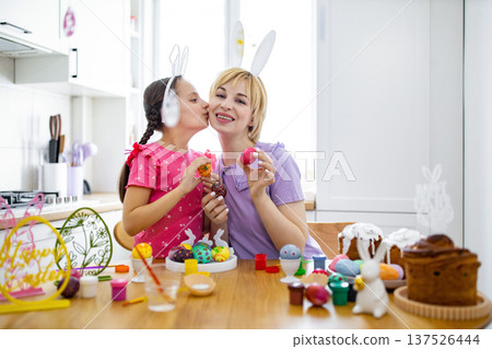 A young girl wearing bunny ears kisses her mother on the cheek while they decorate Easter eggs together in a bright kitchen 137526444