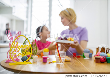 A mother and daughter wearing bunny ears are happily decorating Easter eggs together at a wooden table filled with craft supplies and painted eggs 137526445