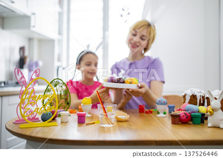 A mother and daughter wearing bunny ears are happily decorating Easter eggs together at a wooden table in a bright kitchen A mother and daughter wearing bunny ears are happily decorating Easter eggs together at a wooden table in a bright kitchen 137526446