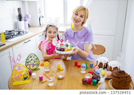 A mother and daughter wearing bunny ears are happily painting and decorating colorful Easter eggs together in a bright kitchen setting A mother and daughter wearing bunny ears are happily painting and decorating colorful Easter eggs together in a bright kitchen setting 137526451
