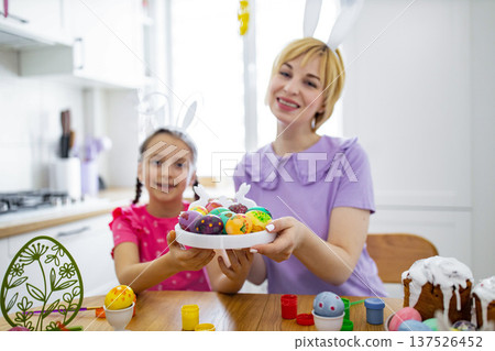 A mother and daughter wearing bunny ears happily display a plate of colorful, hand-painted Easter eggs they have decorated together in a bright kitchen setting 137526452