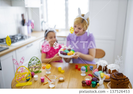 A mother and daughter enjoy decorating Easter eggs together in a bright kitchen, surrounded by festive decorations and cakes A mother and daughter enjoy decorating Easter eggs together in a bright kitchen, surrounded by festive decorations and cakes 137526453